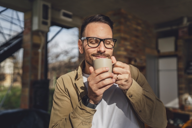 une personne portant des lunettes et une chemise beige tient une tasse à deux mains et semble prendre un verre alors qu'elle se tient debout dans une pièce aux murs en briques apparentes.