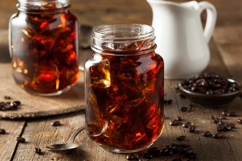 deux pots en verre remplis de café glacé sont posés sur une table en bois à côté d'une cuillère, de grains de café et d'un petit pichet.