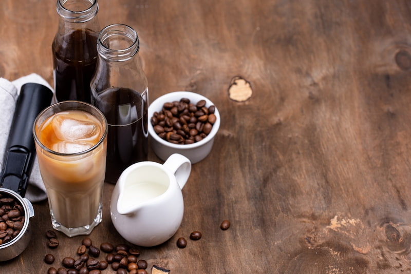 café glacé dans un verre avec du lait, deux bouteilles de café infusé à froid, un petit pichet de lait, des grains de café dans des bols et un presse café sur une table en bois.