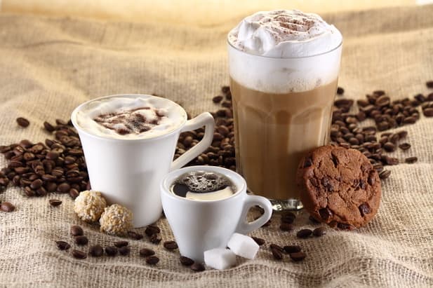 trois tasses avec différentes boissons au café disposées sur une toile de jute, entourées de grains de café, d'un biscuit aux pépites de chocolat, de deux morceaux de sucre et de deux petites truffes.