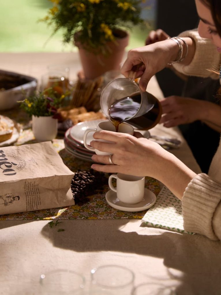 Une personne verse du café d’une carafe en verre dans une tasse blanche posée sur une table dressée avec de la nourriture, un sac de grains de café et une plante en pot.