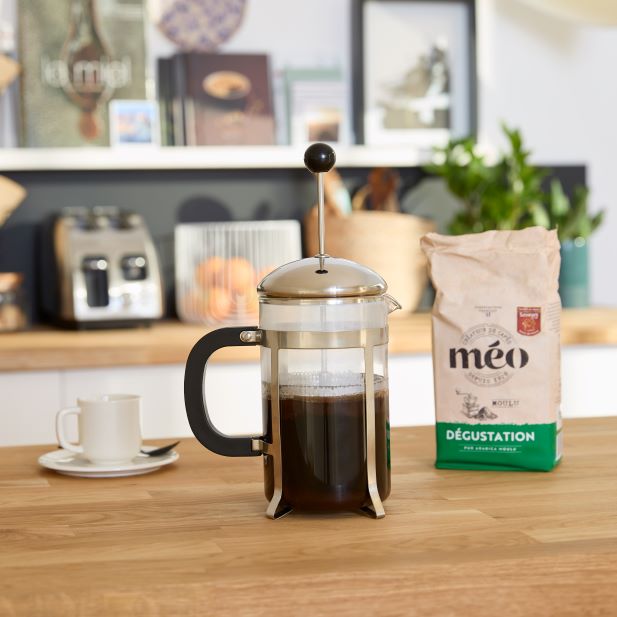 une presse française remplie de café se trouve sur un comptoir de cuisine en bois, à côté d'un sac de grains de café meo. une tasse à café blanche et une soucoupe sont placées en arrière plan.