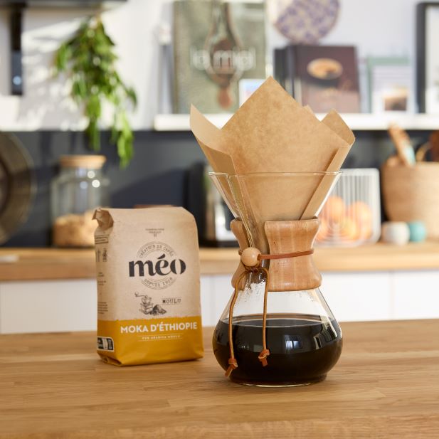 une cafetière chemex avec du café infusé se trouve à côté d'un sac de grains de café moka d'ethiopie sur un comptoir en bois dans une cuisine.