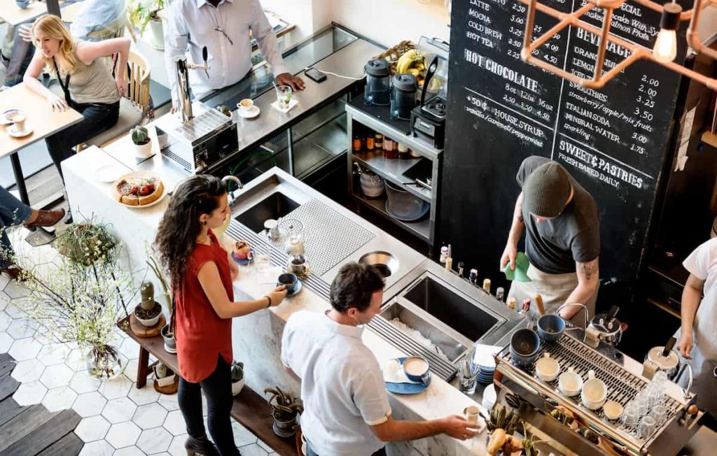 des gens à l’intérieur d’un café animé avec des baristas préparant des boissons derrière le comptoir, pendant que les clients attendent et s’assoient à table. un menu de tableau est visible en arrière plan.