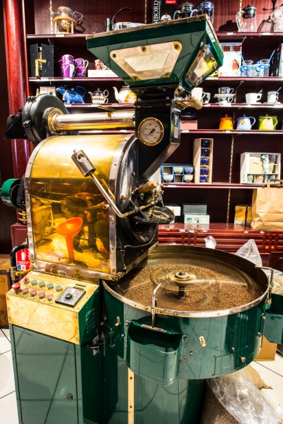 une machine à torréfier le café vert et or avec un panneau de commande dans une vitrine de magasin, entourée de diverses théières et tasses sur des étagères en arrière plan.