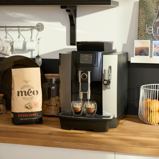 une machine à expresso moderne prépare du café dans deux verres transparents sur un comptoir en bois, à côté d'un sac de grains de café et d'une corbeille de fruits en fil métallique avec des oranges. des étagères avec des tasses sont en arrière plan.