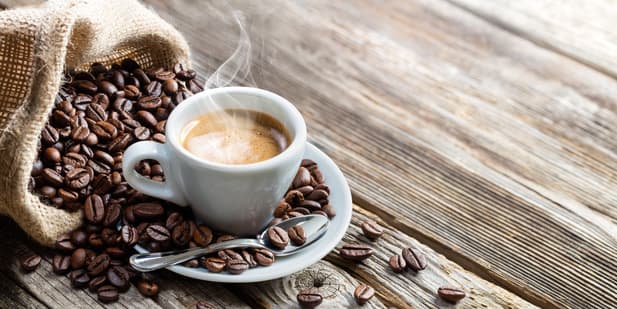 une tasse blanche de café fumant repose sur une soucoupe avec une cuillère, entourée de grains de café épars s'échappant d'un sac en toile de jute sur une surface en bois.