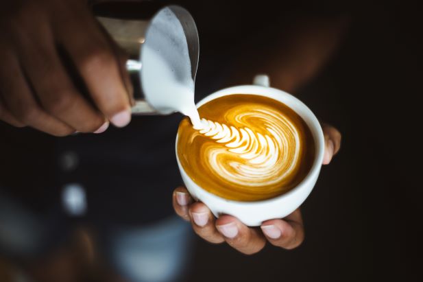 une personne versant de la mousse de lait dans une tasse de café, créant ainsi un latte art avec un motif de feuille.