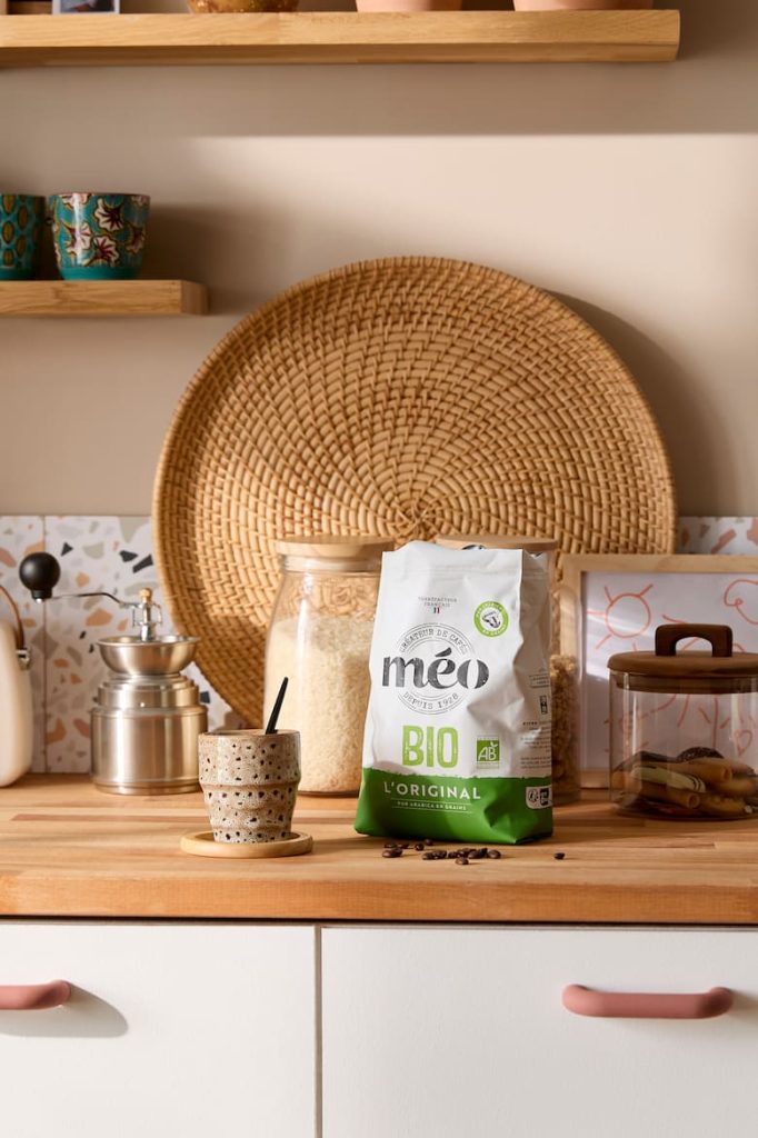 un comptoir de cuisine avec un sac de café méo bio, une tasse en céramique avec une cuillère noire et un pot de riz. des étagères avec des tasses et un panier tressé sont à l'arrière plan.