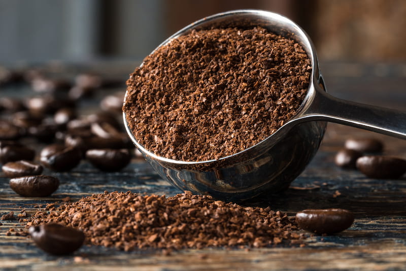 une cuillère en métal remplie de café moulu est renversée, et une partie du café s'écoule sur une surface en bois. des grains de café sont éparpillés à l'arrière plan.