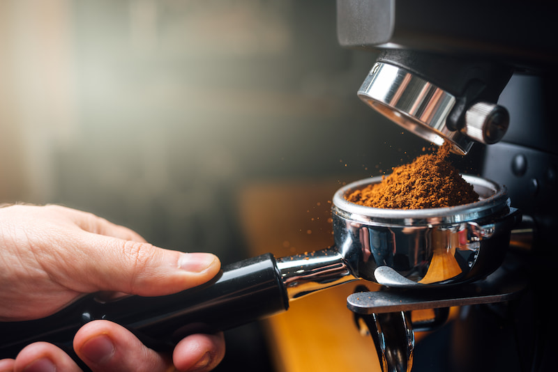 une main tient un porte filtre recevant du café fraîchement moulu provenant d'un moulin électrique.