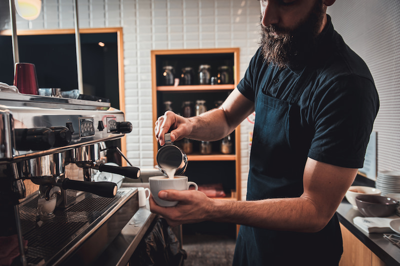 barista avec une barbe versant du lait dans une tasse de café à partir d'un pichet en métal à côté d'une machine à expresso dans un café.