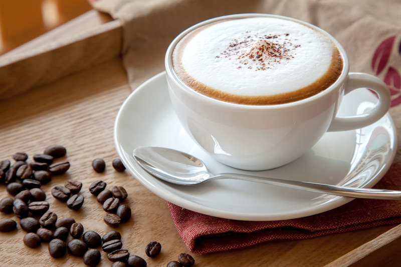 un cappuccino dans une tasse blanche sur une soucoupe avec une cuillère, posé sur un plateau en bois. des grains de café sont dispersés à proximité sur un tissu rouge.