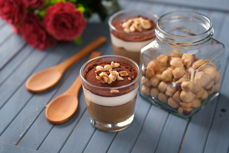 deux verres de dessert au chocolat en couches garnis de noix sont posés sur une table en bois bleu à côté de deux cuillères en bois et d'un pot de noisettes avec des fleurs rouges en arrière plan.