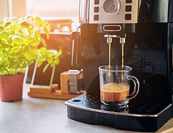 machine à expresso préparant du café dans une tasse en verre transparent, avec une plante de basilic et des ustensiles de cuisine en arrière plan.